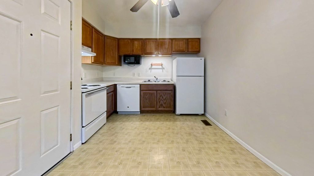 Apartment kitchen with wood cabinets, white appliances, and open floor space at Emerald Pointe Senior Living.